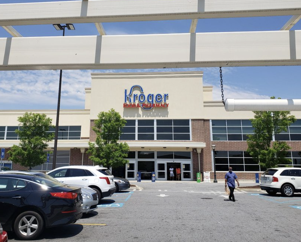 The exterior of a Kroger grocery store with parked cars in front, a person walking towards the entrance, and trees on either side under a partly cloudy sky.