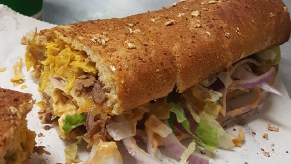 A close-up of a sub sandwich on oat bread, filled with shredded lettuce, red onions, steak, crushed chips, and a creamy orange sauce, resting on white parchment paper.