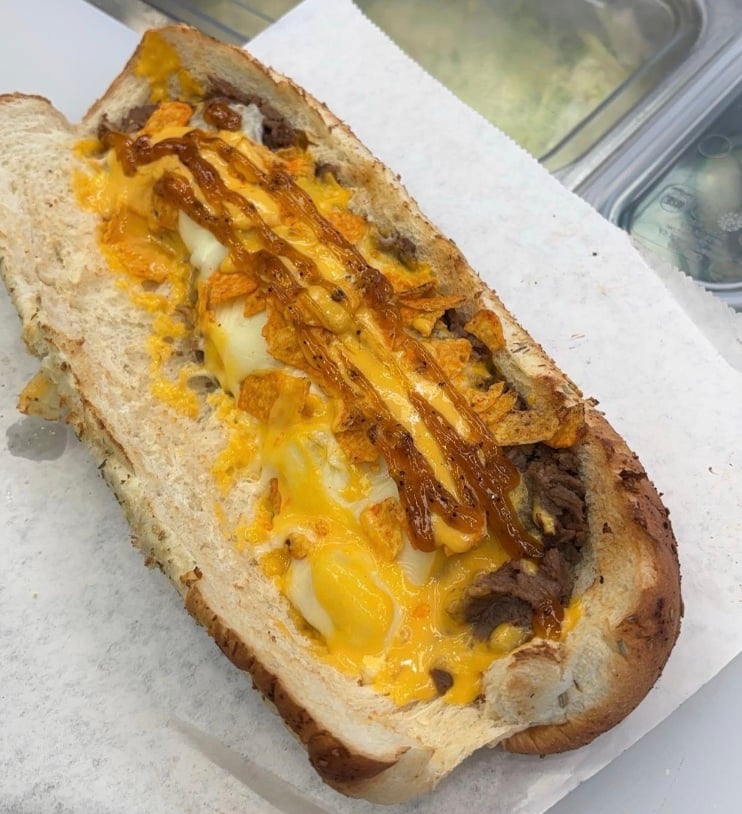 A hoagie roll filled with melted cheese, steak, crushed chips, and a drizzle of sauce, served on a sheet of white parchment paper.
