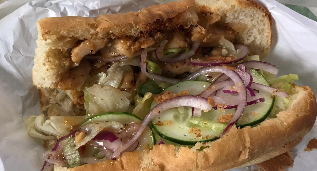 A close-up of a sandwich with shredded lettuce, cucumber slices, red onion, grilled chicken, and sauce in a partially sliced hoagie roll, placed on white paper.