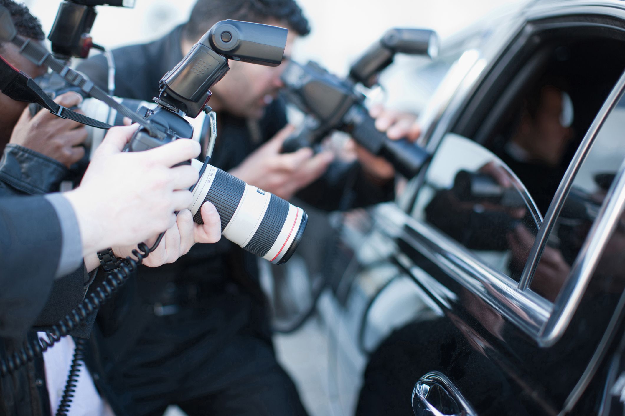 Three paparazzi members with cameras taking photos of someone inside a the back seat of a black car with the window down, left side of the car, blurred background
