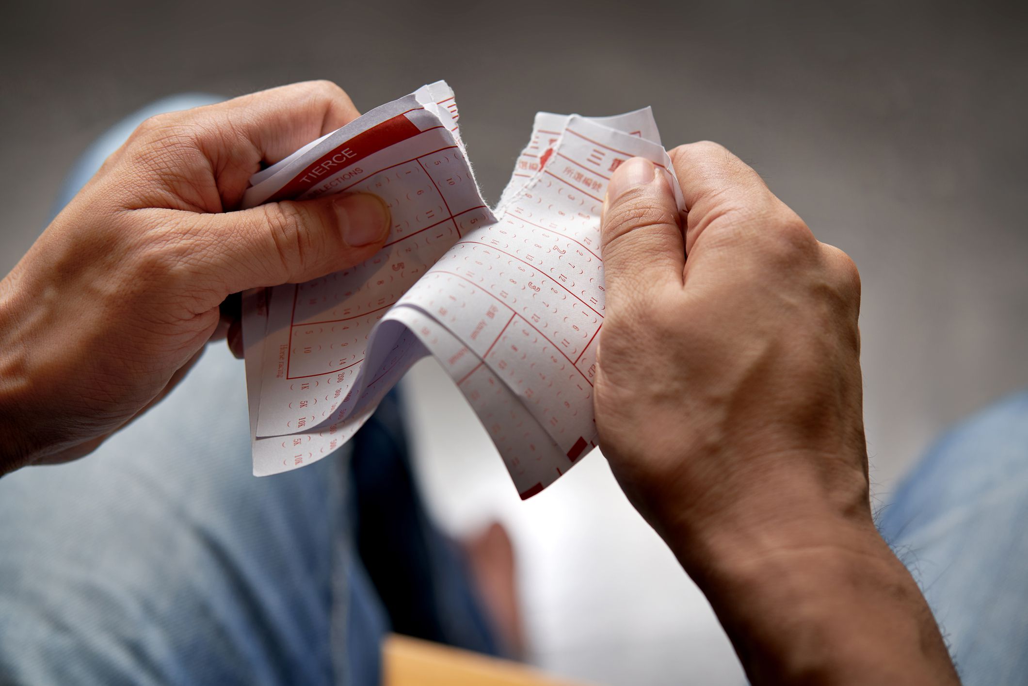 Two hands tearing horse racing tickets in half, legs in jeans and grey floor blurred in the background