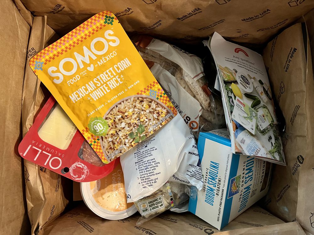 An open cardboard box filled with various packaged foods, including SOMOS Mexican street corn rice, sliced cheese, yogurt cups, and other assorted grocery items.