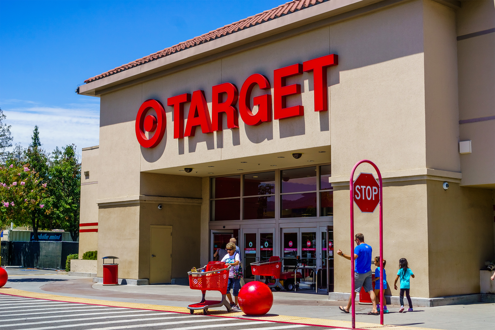 People walk with shopping carts outside a Target store on a sunny day. Large red Target sign is above the entrance; red bollards and a stop sign are visible in front of the building.