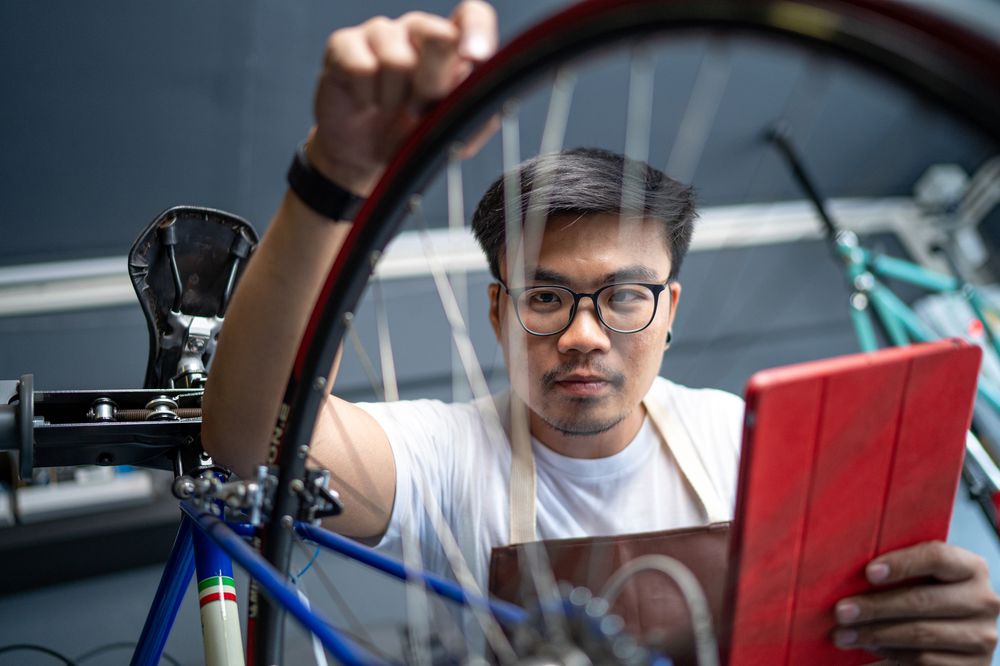 A man wearing glasses and an apron examines a bicycle wheel while holding a red tablet, focusing intently as he works in a bike repair shop.