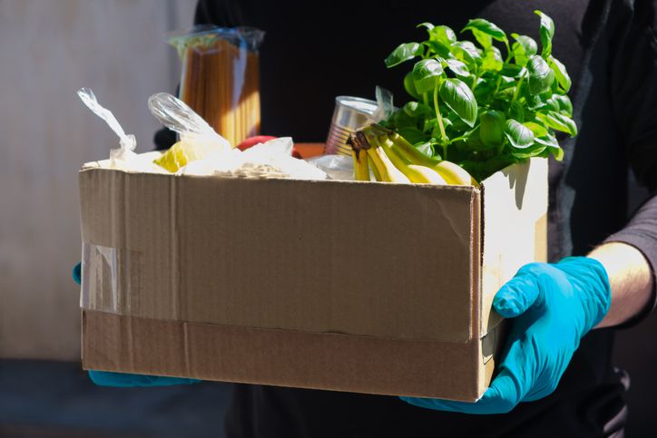 A person wearing blue gloves holds a cardboard box filled with groceries, including bananas, canned food, pasta, a bunch of basil, and other food items.