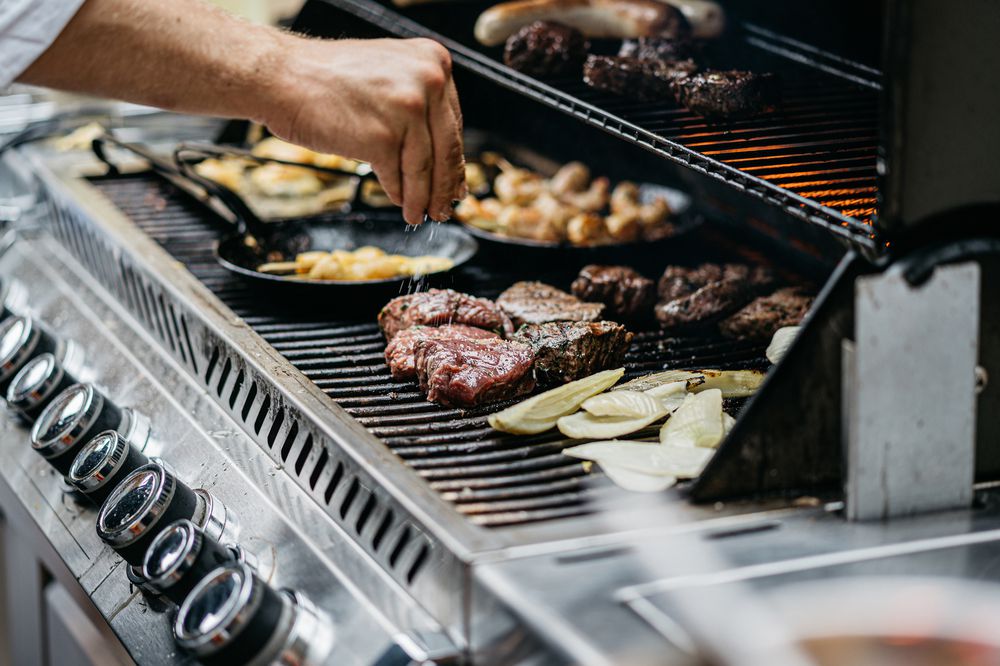 A person sprinkles seasoning onto steaks and onions grilling on a barbecue, with various meats and vegetables cooking on both levels of the grill.