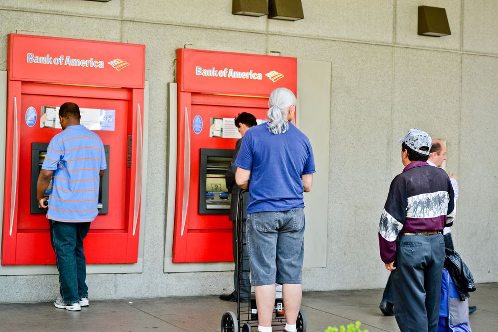 Four people stand in line at two red Bank of America ATMs outside a building; one man uses an ATM, while others wait, including a man with a ponytail and cart, and another wearing a patterned jacket and cap.