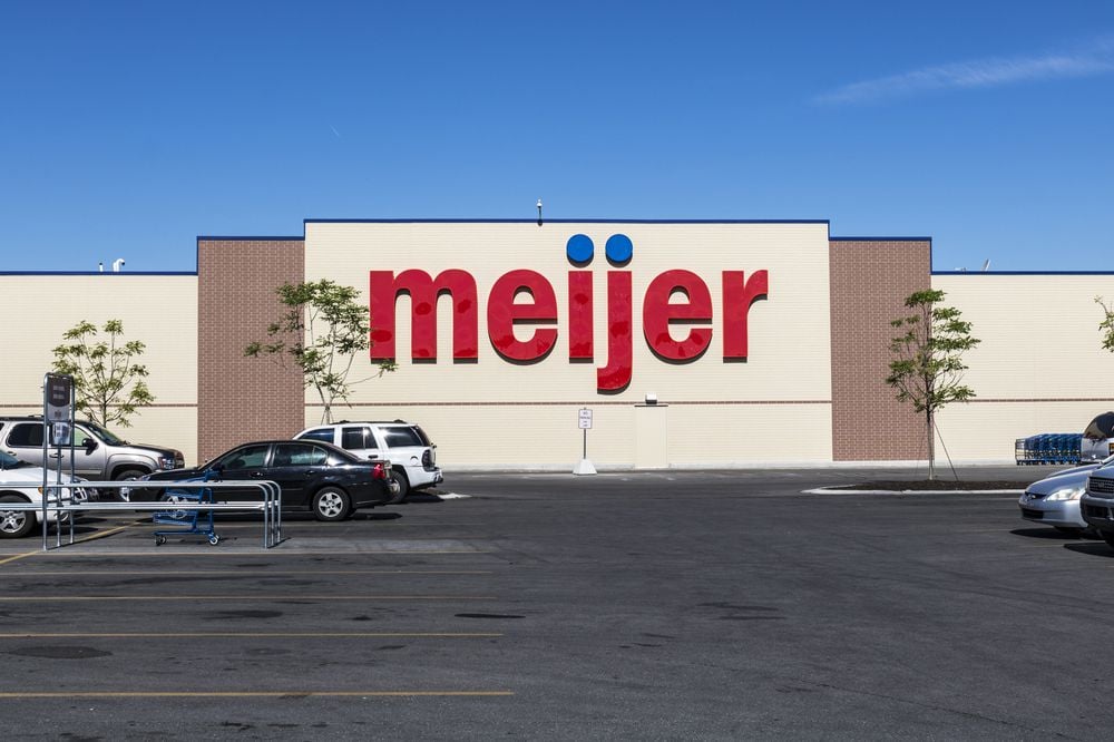 The front of a Meijer store with large red letters on the building, cars parked outside, shopping carts in the lot, and a clear blue sky overhead.