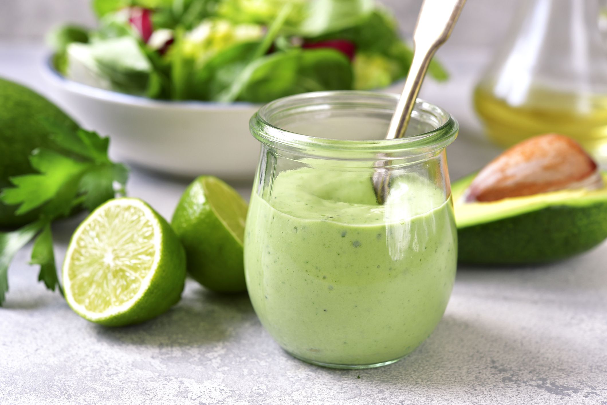 Homemade avocado yogurt dressing in a vintage glass jar with ingredients for making on a light slate, stone or concrete background.
