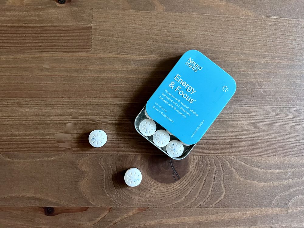 A blue tin labeled “Energy & Focus” contains mints, with four mints inside and three more scattered on a wooden surface. The mints are round with a pattern resembling a flower on top.