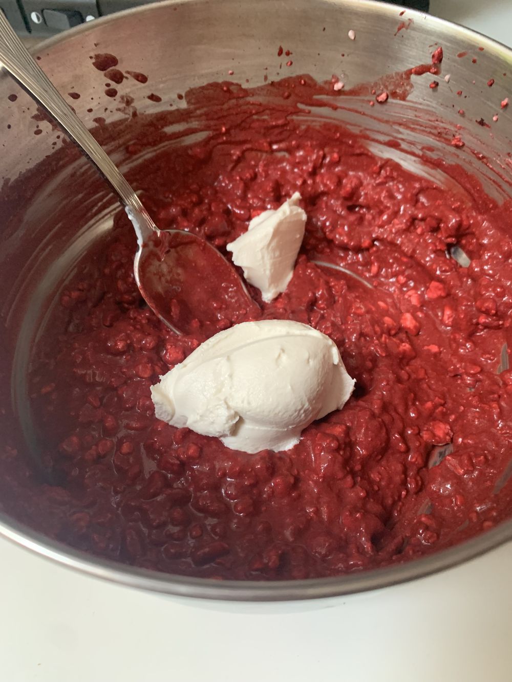 A metal bowl filled with a thick, red batter mixed with small chunks, topped with a spoonful of white cream cheese. A metal spoon rests inside the bowl.