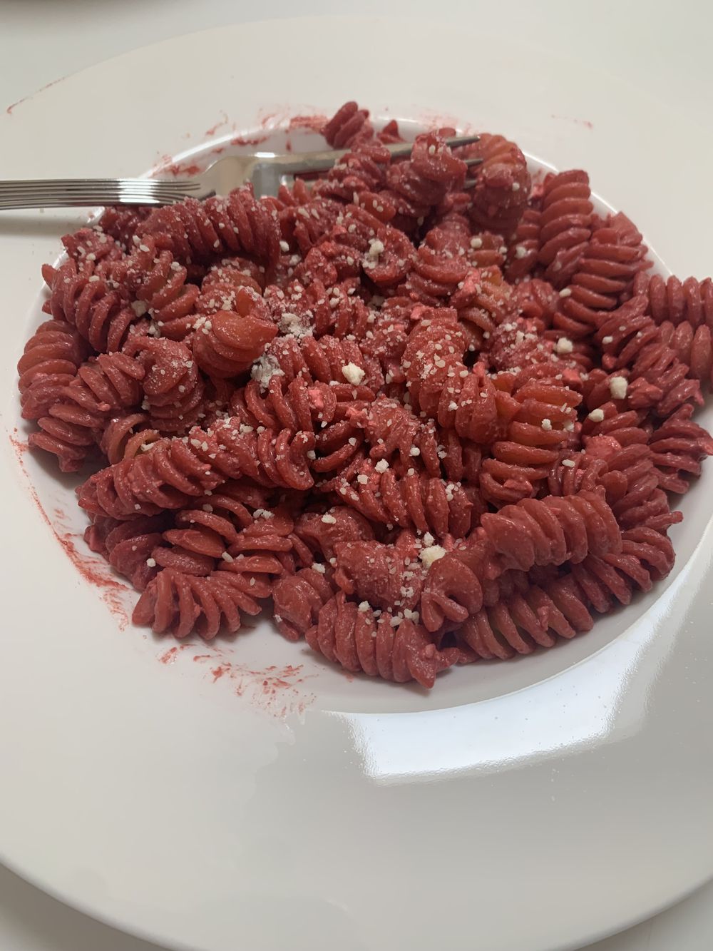 A white plate with red rotini pasta, topped with grated cheese, and a fork resting on the edge of the plate. The pasta appears to be beet or tomato infused.