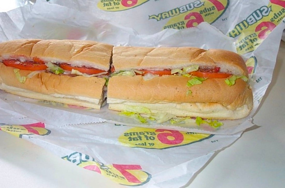 A sub sandwich with lettuce, tomato, and mayonnaise sits on a sheet of branded Subway sandwich paper, cut in half and placed on a white surface.