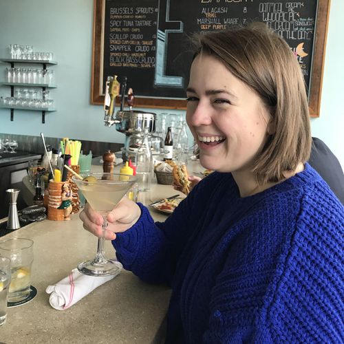 A woman in a blue sweater smiles while holding a martini glass at a bar, with drinks, glassware, and a chalkboard menu visible in the background.
