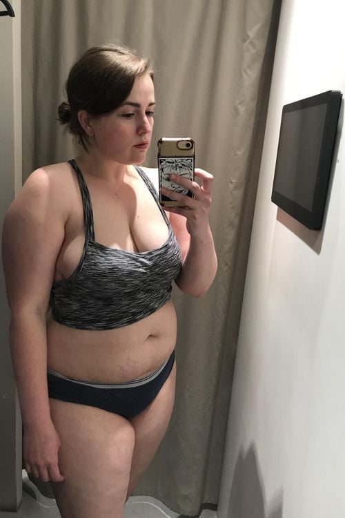 A woman wearing a gray sports bra and dark underwear takes a mirror selfie in a fitting room with beige curtains and a mounted screen on the wall.