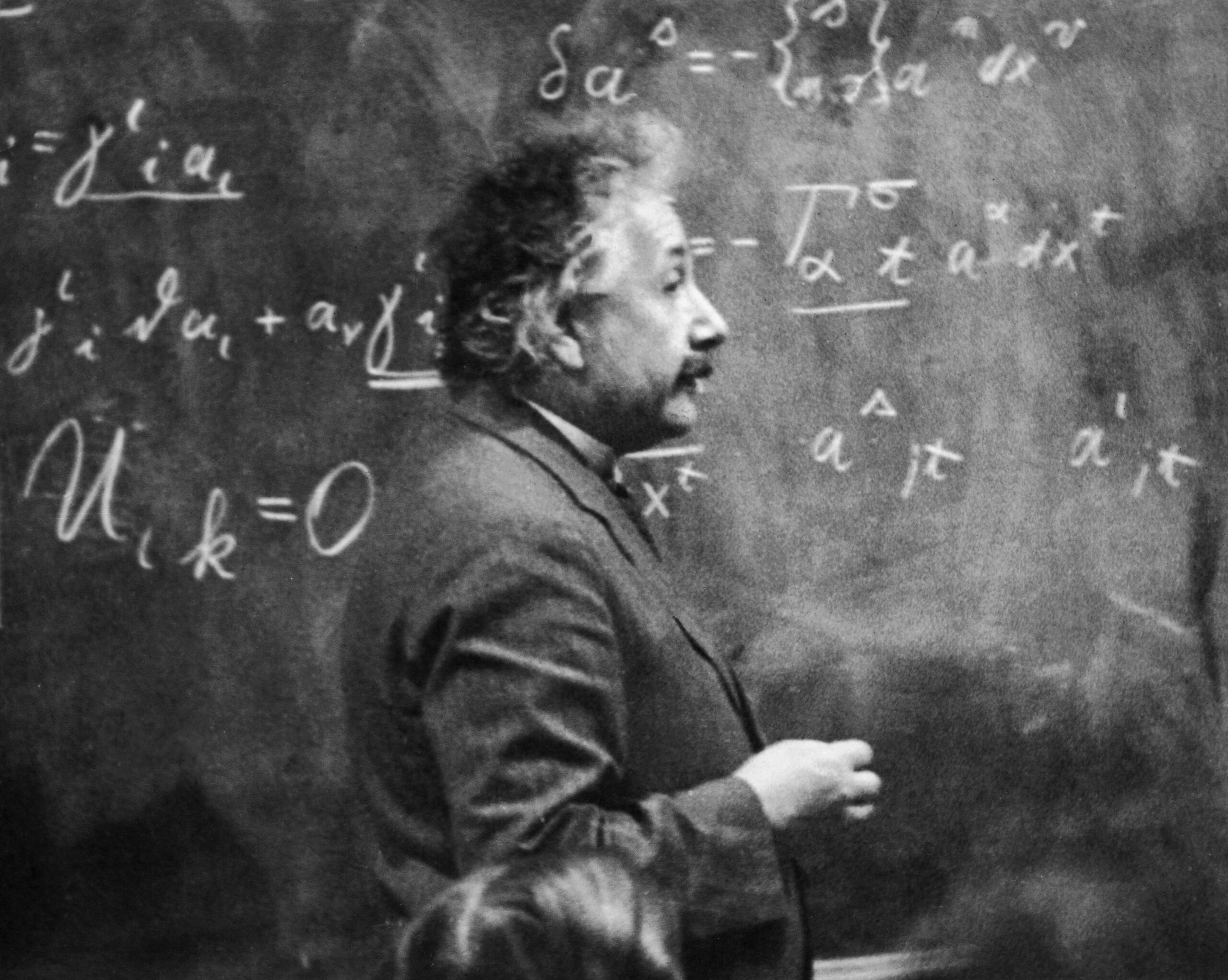 Sideview of Albert Einstein standing beside a blackboard with chalk-marked mathematical calculations written across it, as he is talking to someone sitting off to the side, black and white photograph