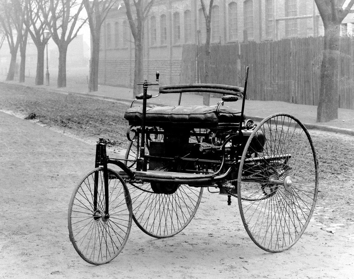 Benz Patent-Motorwagen Nr. 1, black and white image, against a background of an old building and a tree-lined road