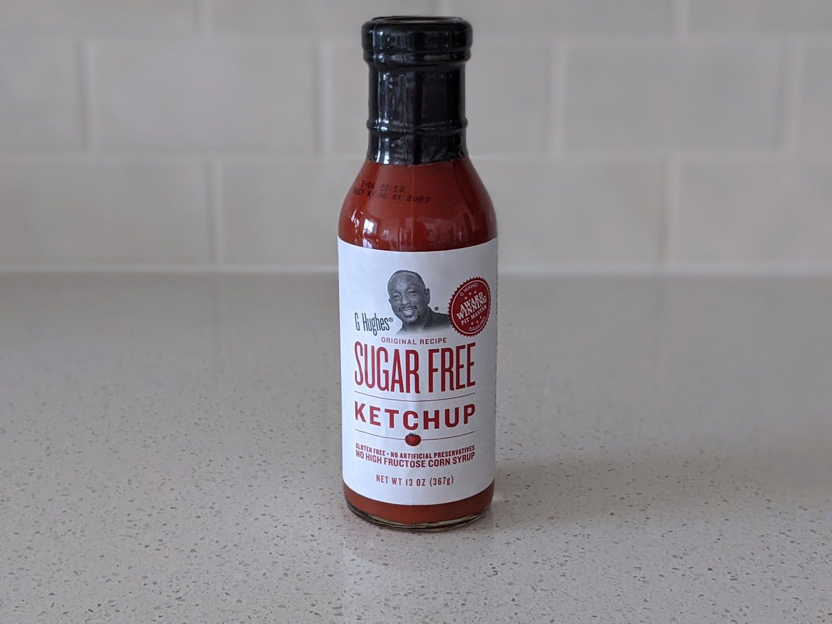 A bottle of G. Hughes Original Recipe Sugar-Free Ketchup on a speckled ivory kitchen countertop, light beige tile wall in the background