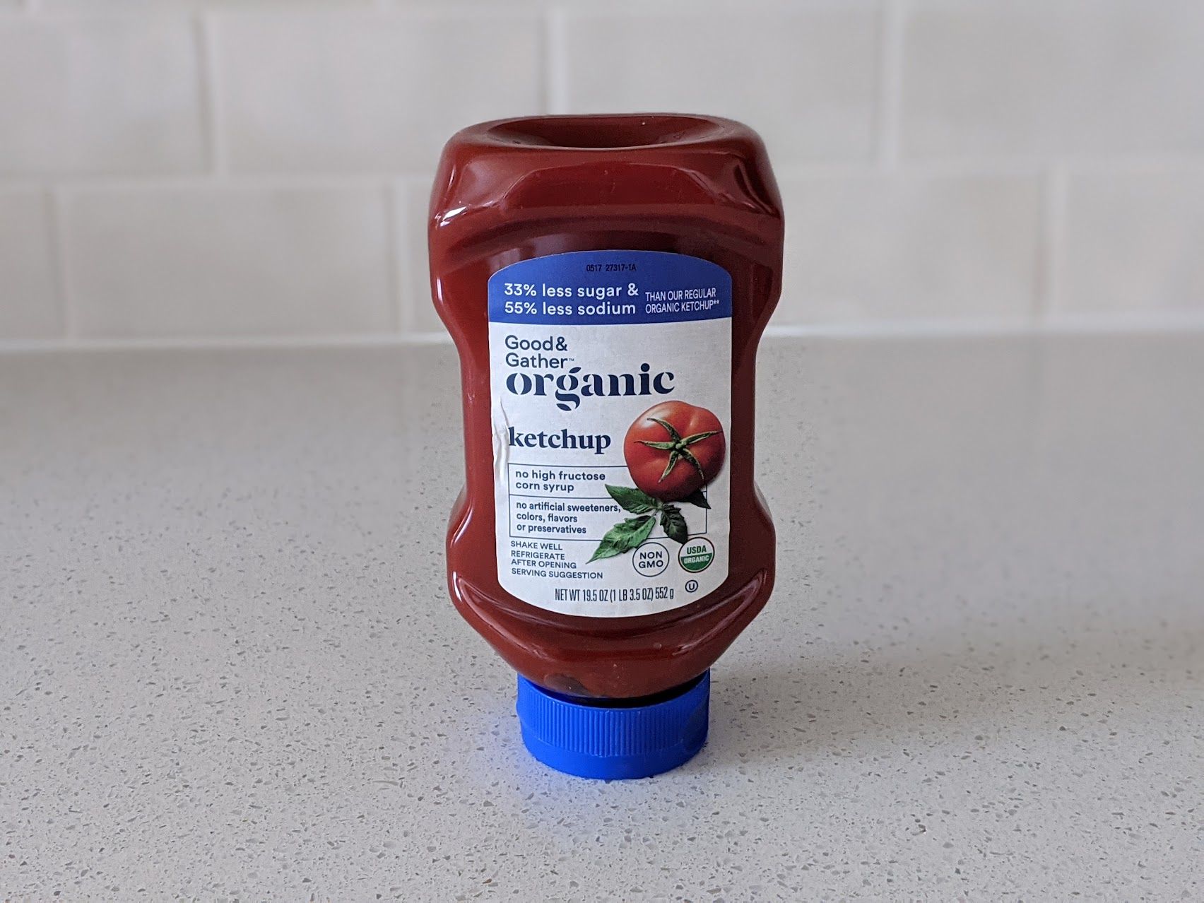 A bottle of Good & Gather Organic Ketchup Reduced Sugar and Sodium on a speckled ivory kitchen countertop, light beige tile wall in the background