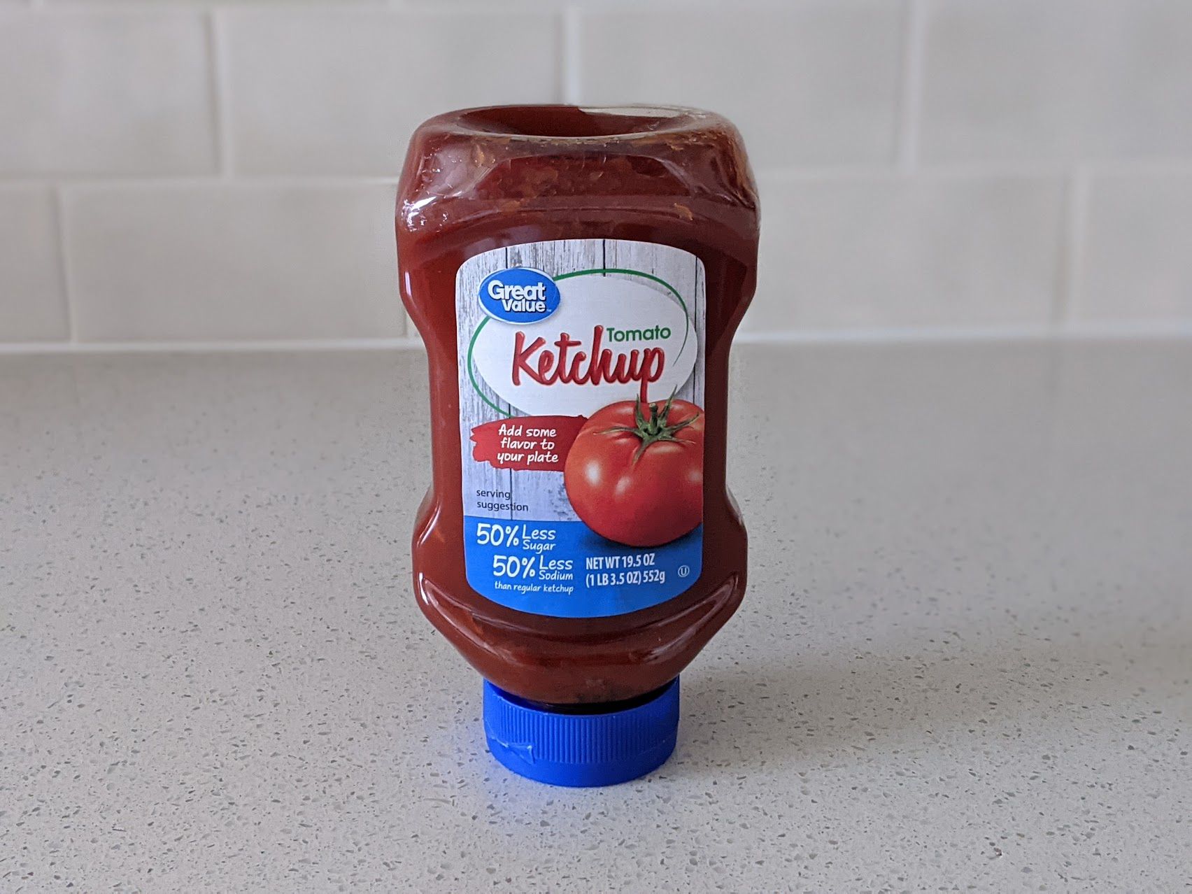 A bottle of Great Value 50% Less Sugar, 50% Less Sodium Ketchup on a speckled ivory kitchen countertop, light beige tile wall in the background