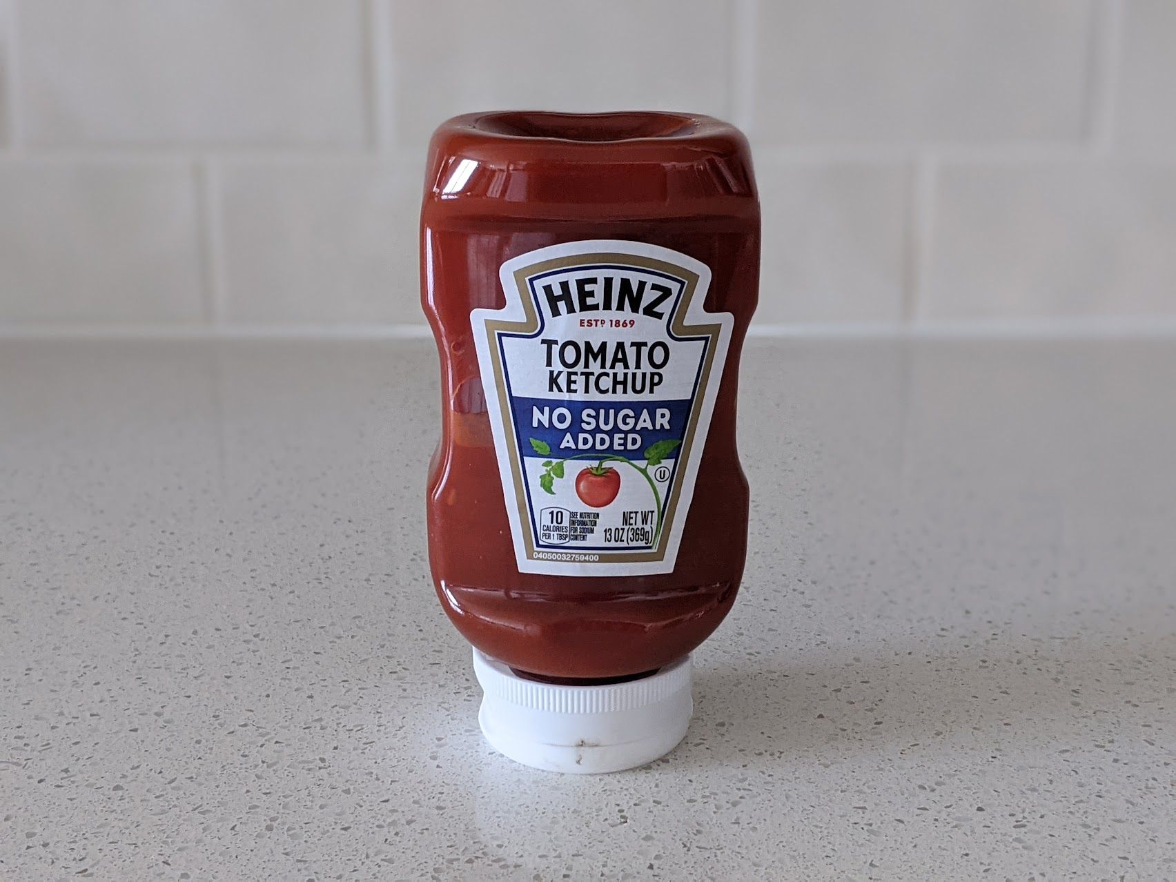 A bottle of Heinz No Sugar Added Ketchup on a speckled ivory kitchen countertop, light beige tile wall in the background