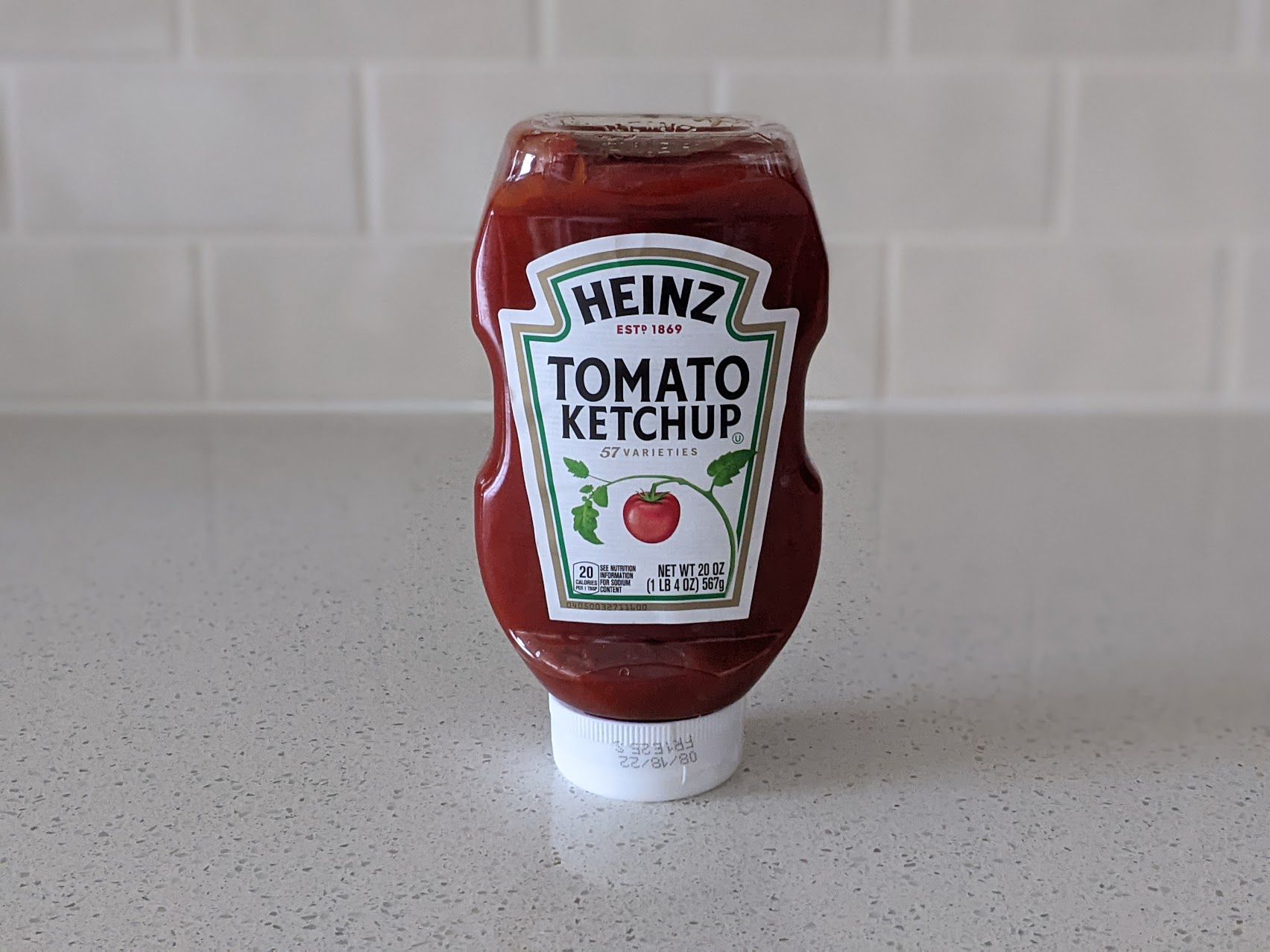 A bottle of Heinz Tomato Ketchup on a speckled ivory kitchen countertop, light beige tile wall in the background