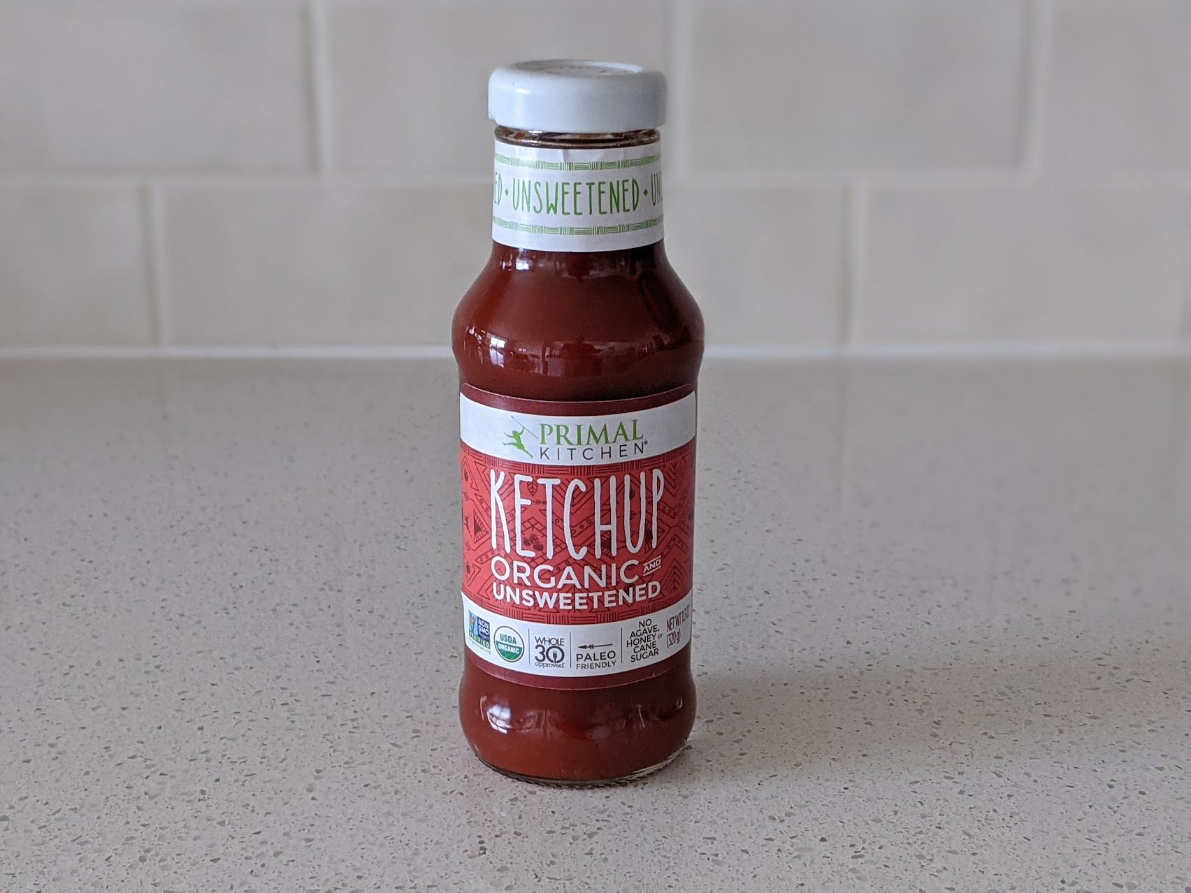 A bottle of Primal Kitchen Organic and Unsweetened Ketchup on a speckled ivory kitchen countertop, light beige tile wall in the background