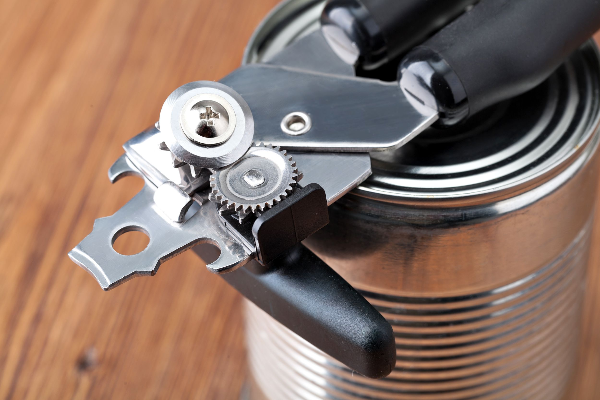 Closeup of manual can opener laid on top of plain closed can, no wrapper on it, selective focus, blurred background of wooden veneer table