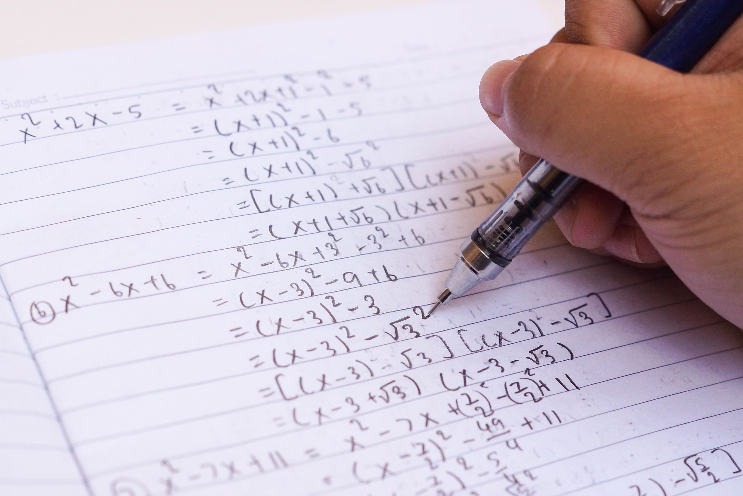 Closeup of hand holding a mechanical pencil, writing algebra in a notebook of college lined paper, slight diagonal view with beige table in the top left corner