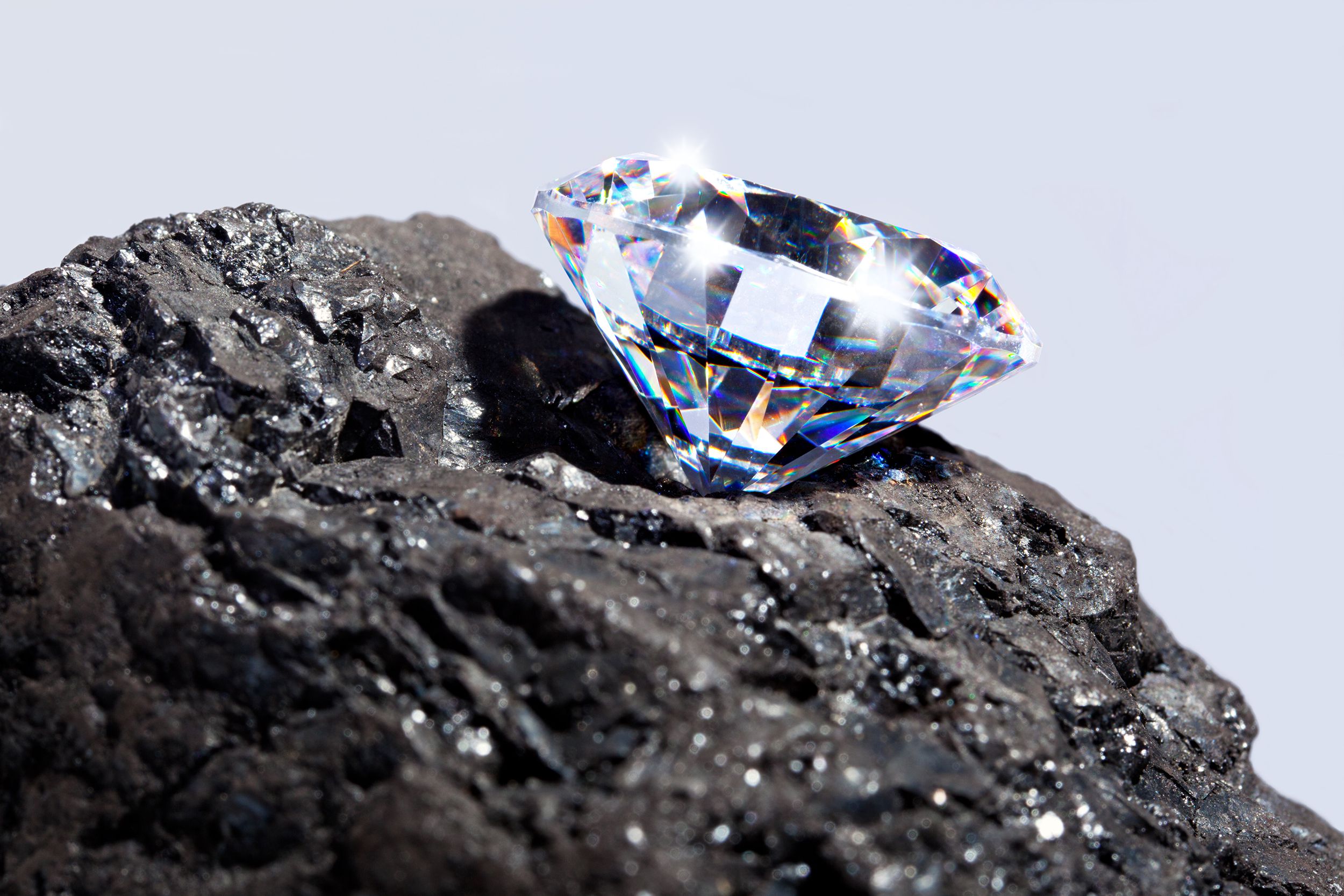 Closeup of a single round cut diamond, colorful sparkling in the light, on a hunk of coal, against a light grey background