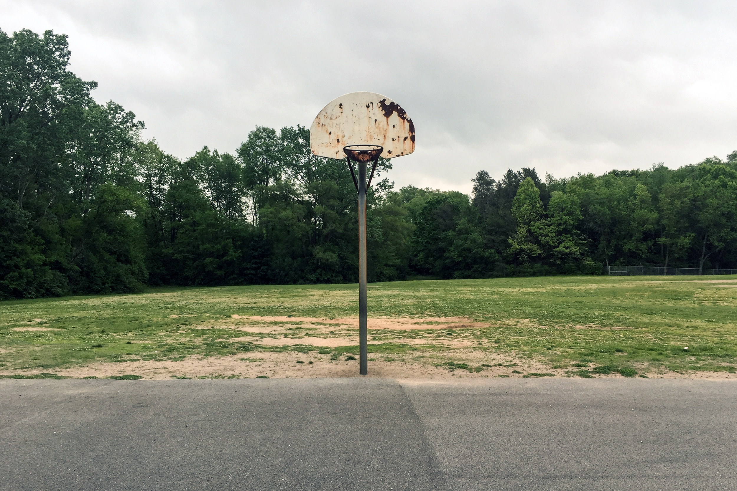 Dilapidated basketball hoop with a cement area in the foreground, grass and a line of trees in the background, cloudy sky