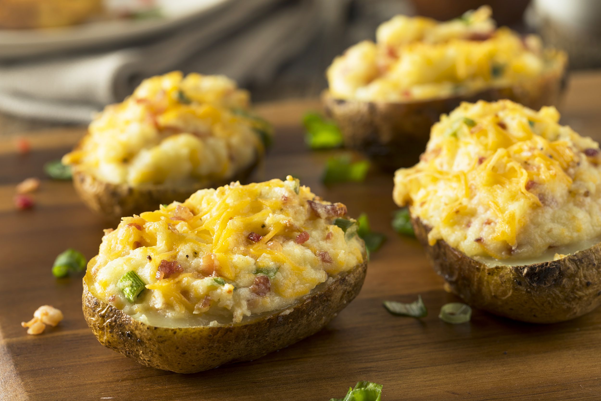 Four loaded potatoes on an artisanal wooden board, selective focus, surrounded by herbs with a blurred background of napkins and another food dish