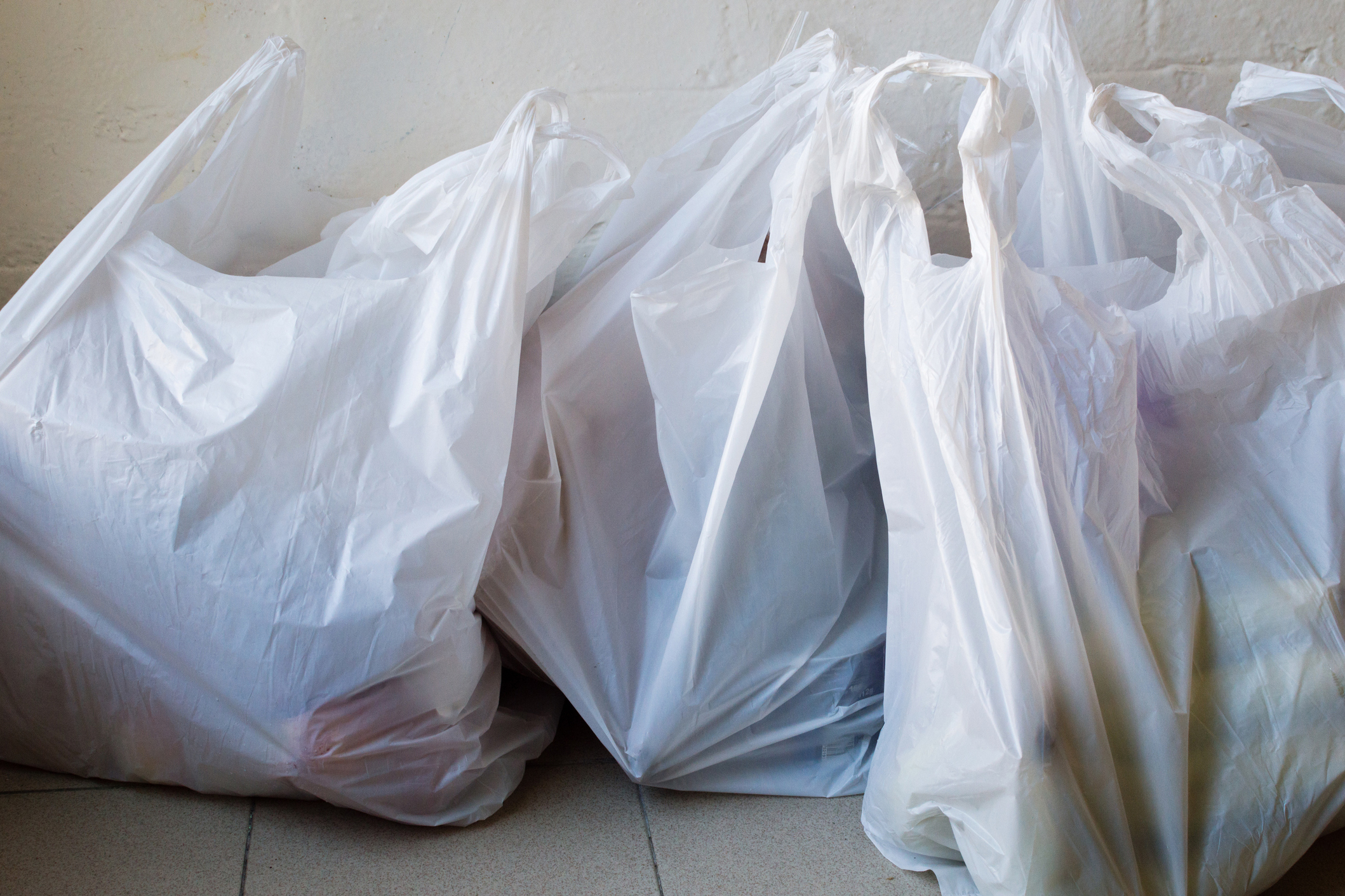 Four plain white plastic bags filled with groceries on beige tiled floor, beige wall in the background