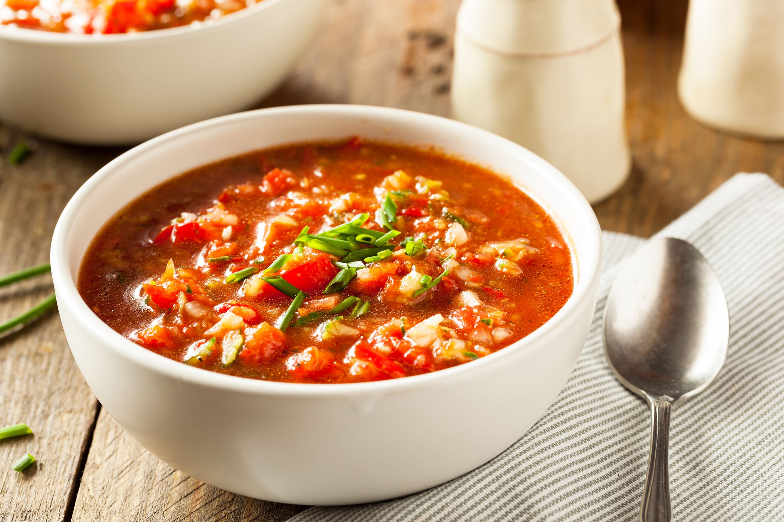 Gazpacho in a white porcelain bowl with a blue-lined white napkin and spoon on a rustic wooden table with another bowl of gazpacho and white salt and pepper shakers blurred in the background