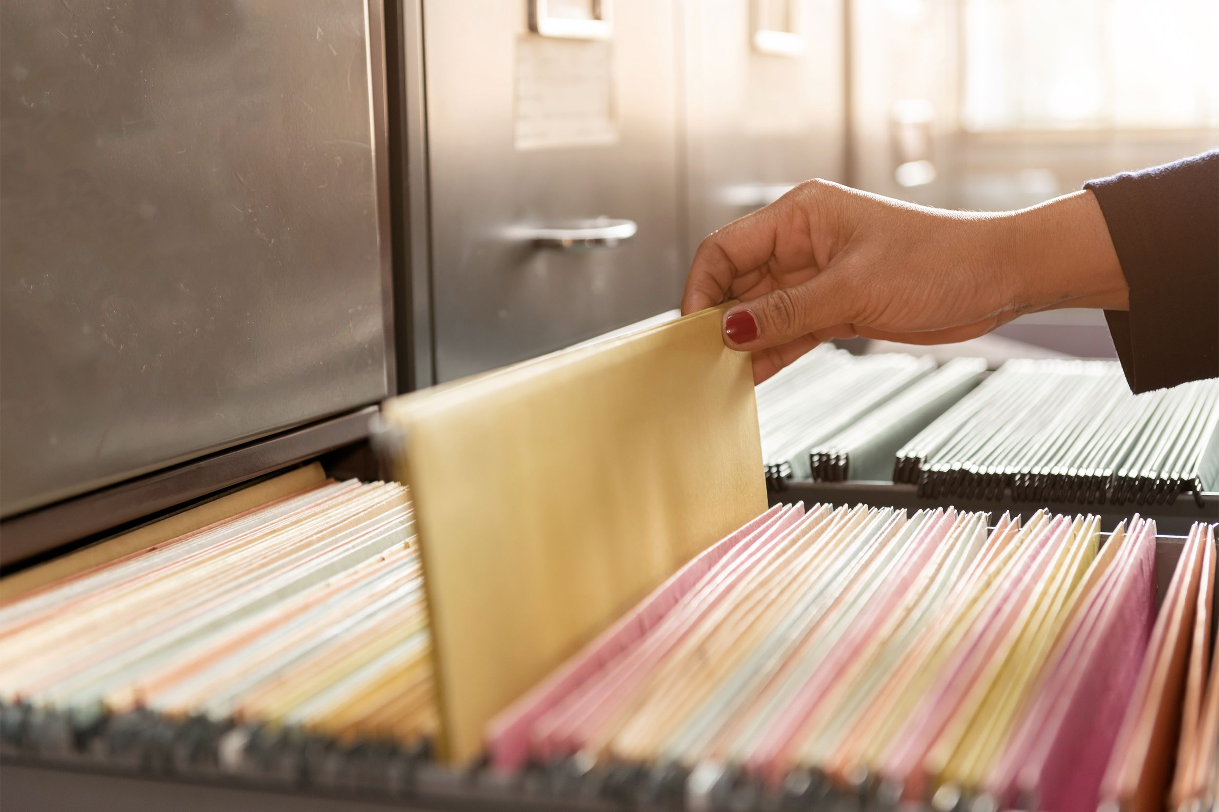 Female hand pulling a hanging file from a file filled drawer of a filing cabinet in the foreground, by an adjacent opened file filled drawer, surrounded by a top row of closed drawers of filing cabinets