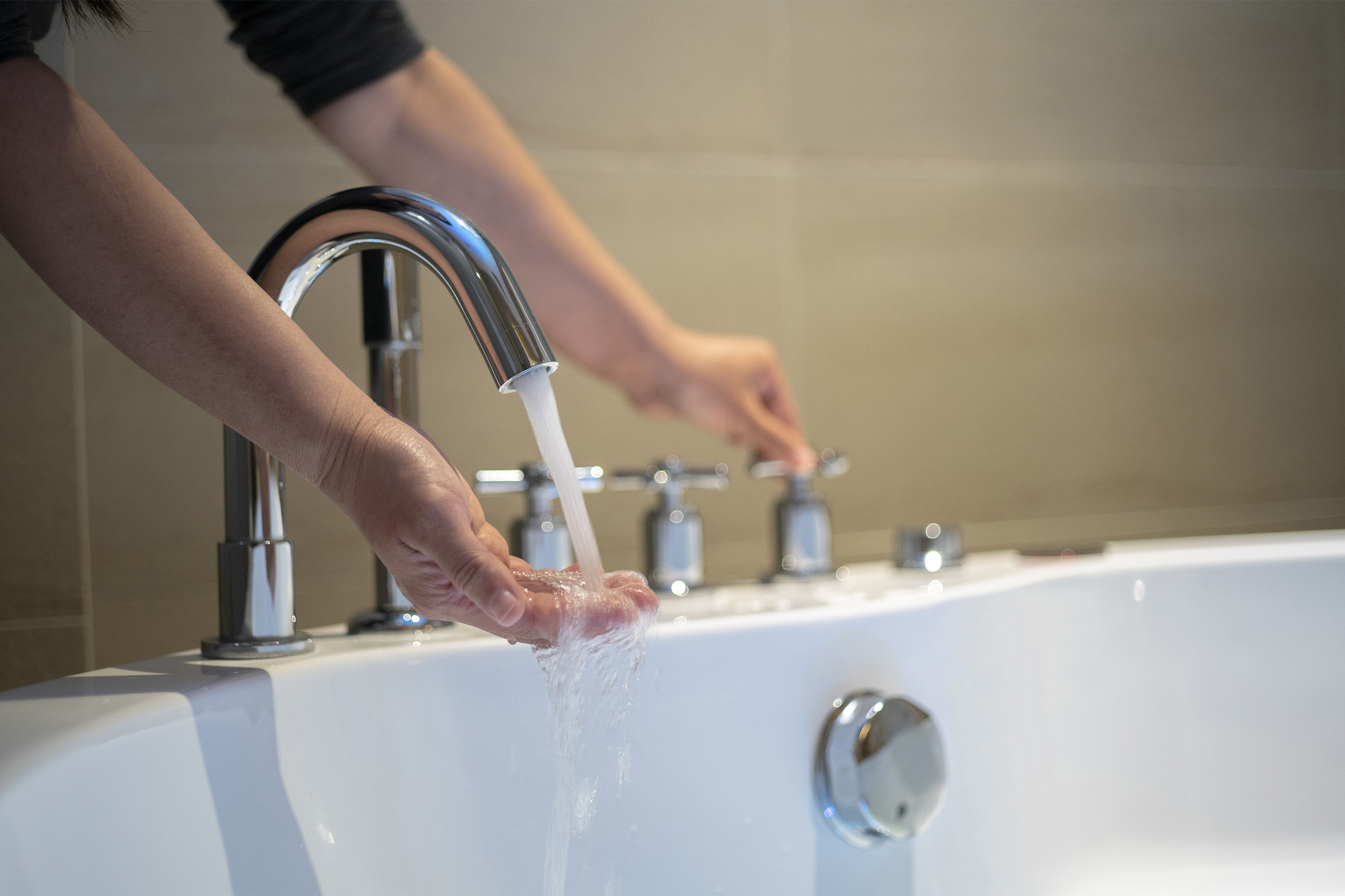 Hand turning on water of a white bathtub surrounded by tile, selective focus of hand feeling water coming out of faucet, blurred background of beige bathroom tiled walls