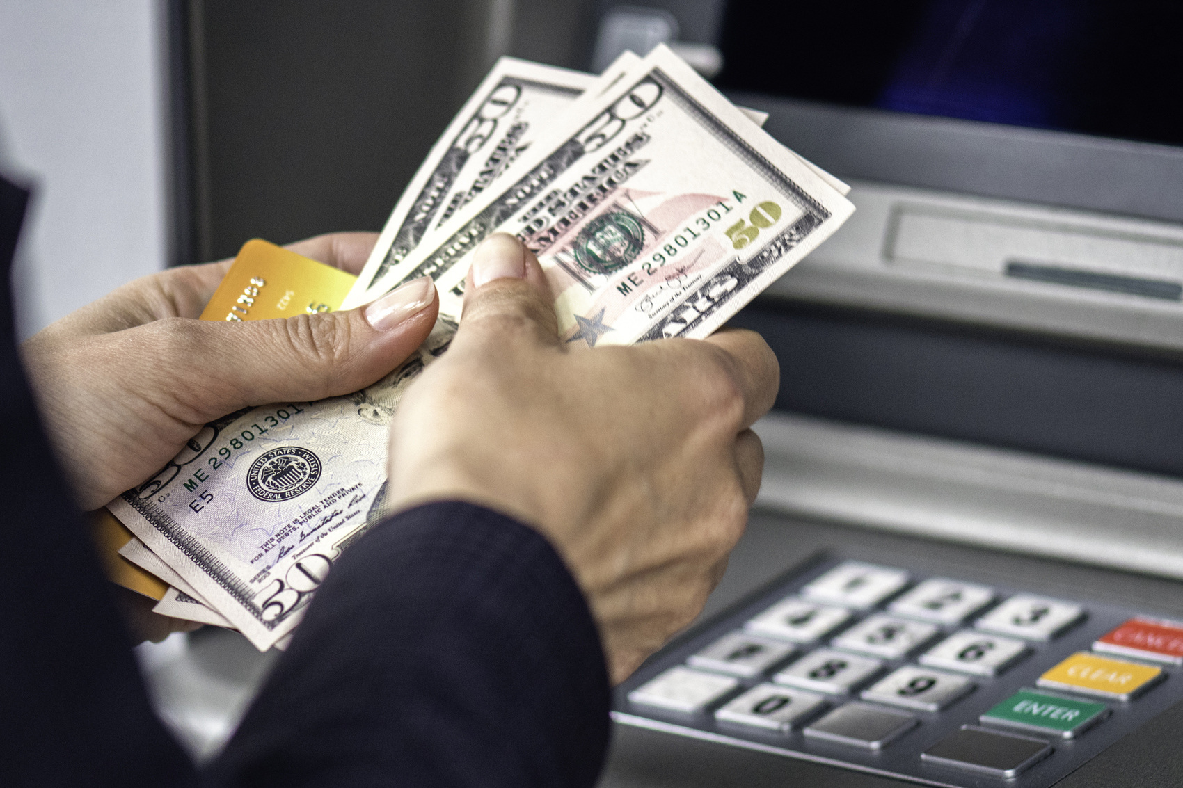 Hands holding cash in three $50 dollar bills, just received from a bank ATM, selective focus, ATM blurred in the background