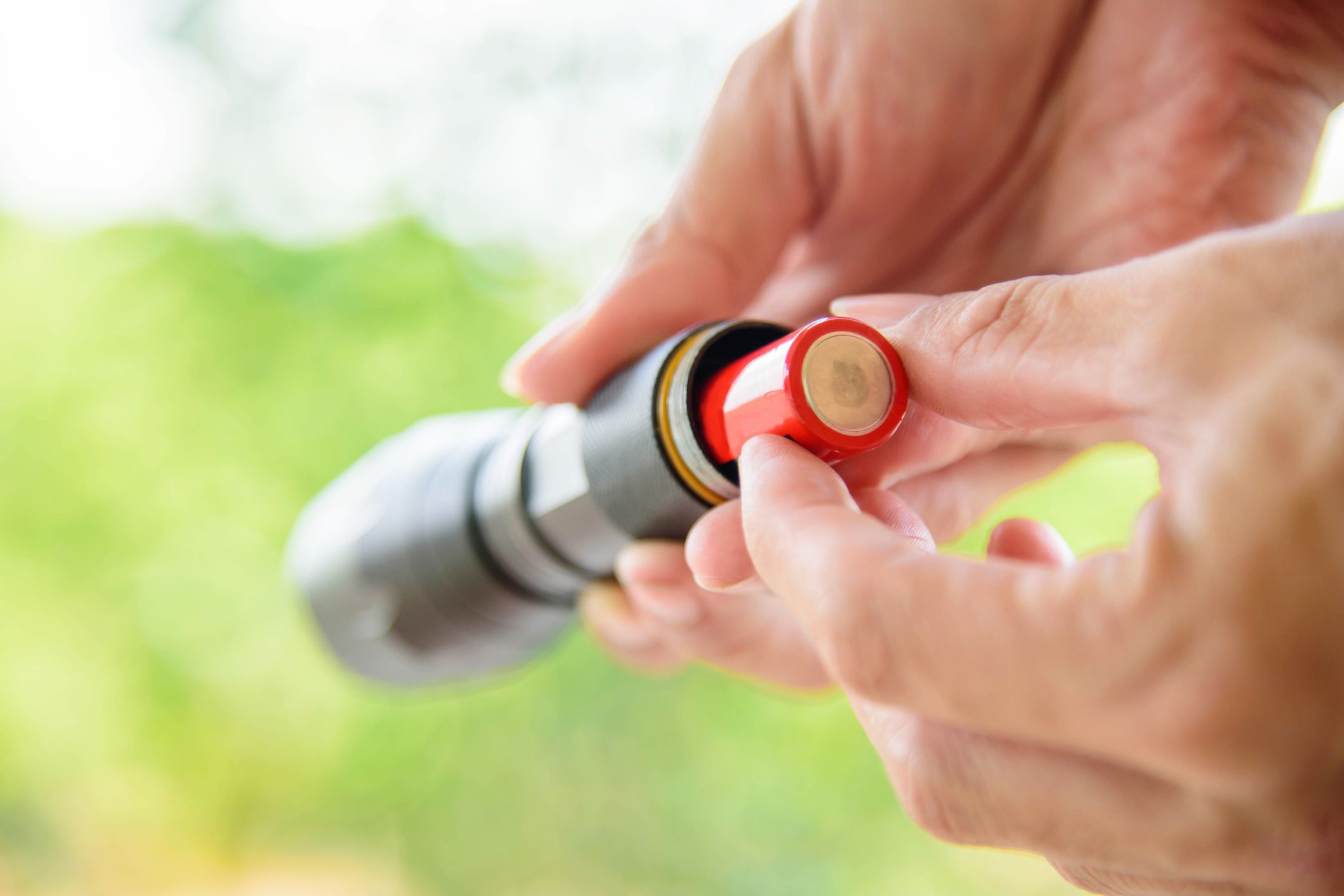 Hands putting AA batteries into a grey flashlight, selective focus, blurred background of green and white outdoors