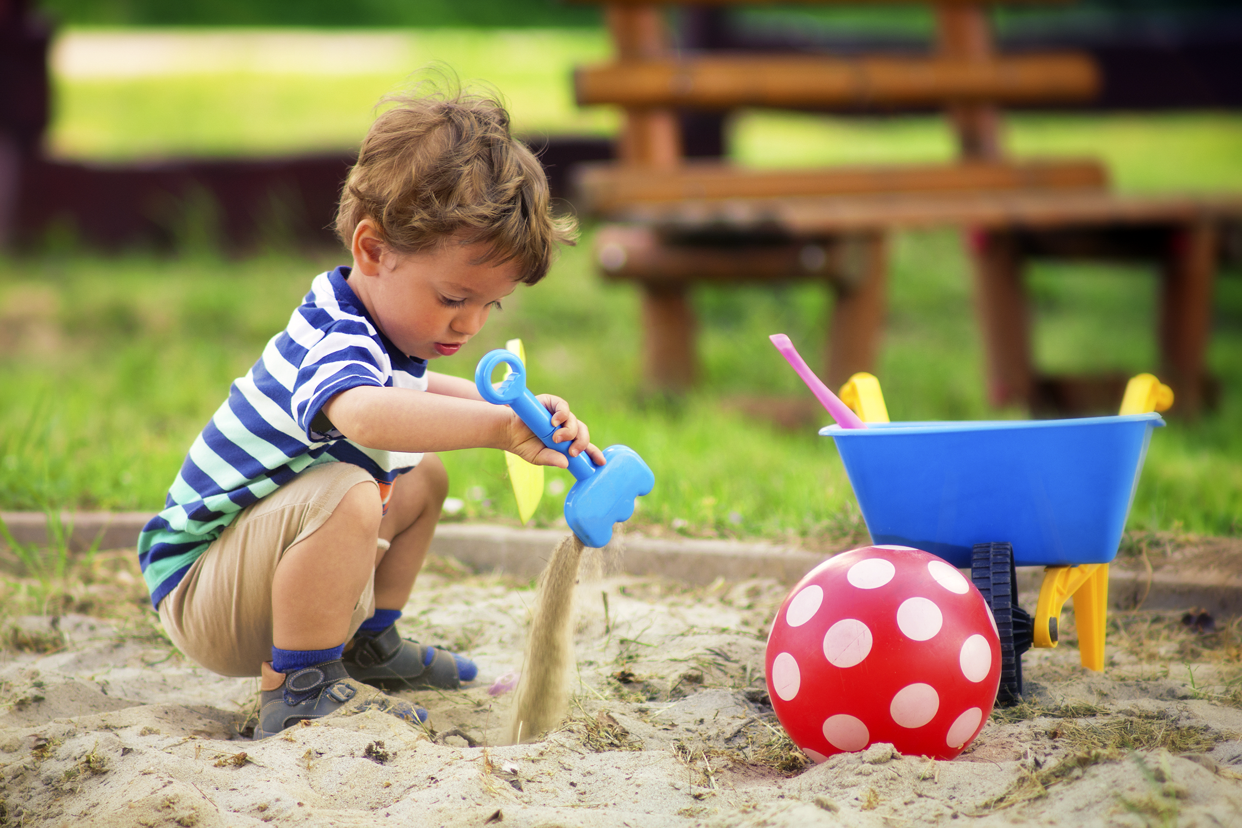 Little boy playing in a sandbox, selective focus, he is pouring sand from a shovel while squatting, in a playground, grass and a wooden bench in the background