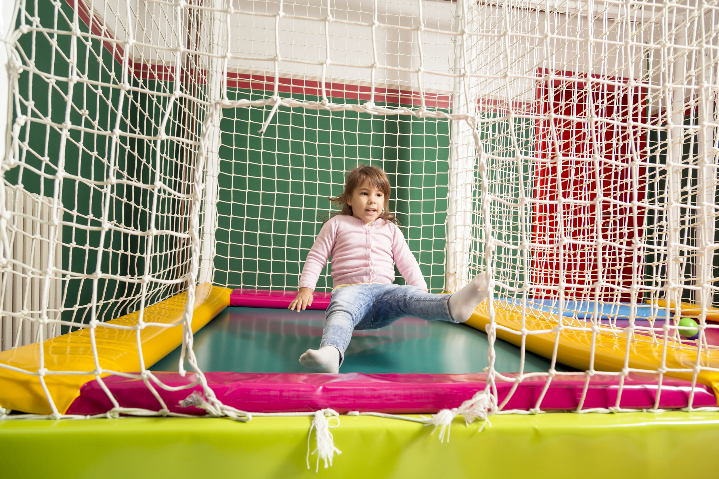 Little girl sitting in the air while playing on an indoor trampoline, surrounded by a robe fence, as seen from outside, walls and another trampoline on the right