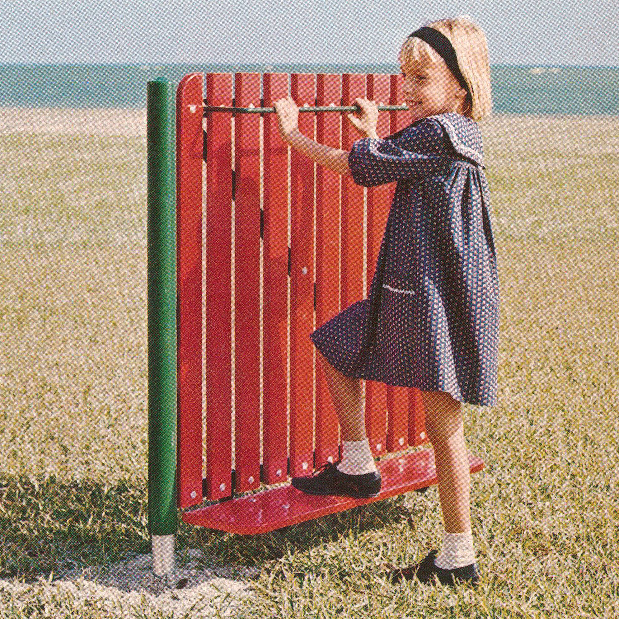 Little girl smiling while on a red metal swinging gate, on grass, she is facing towards the left, vintage, the beach and ocean in the background