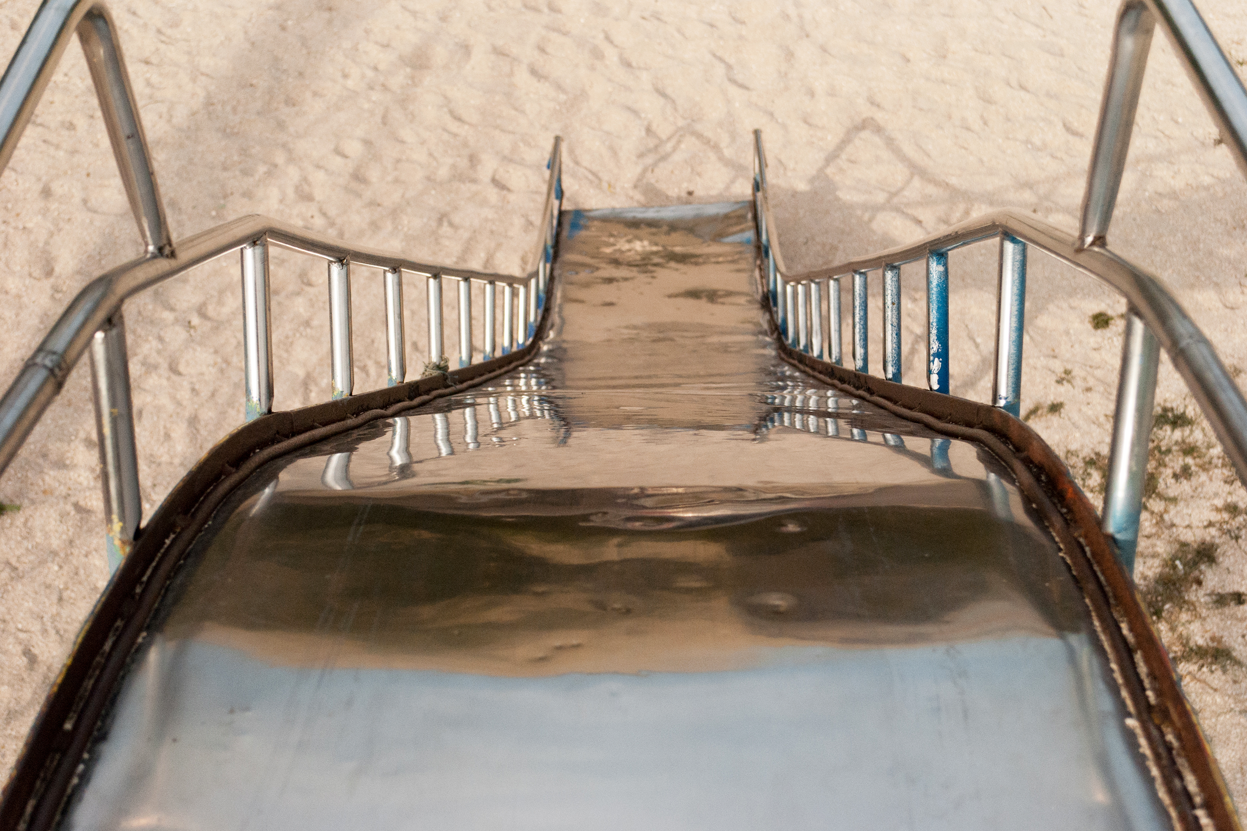 Metal slide as viewed from above looking down it, metal handrails on both sides, sand on the bottom, on a playground