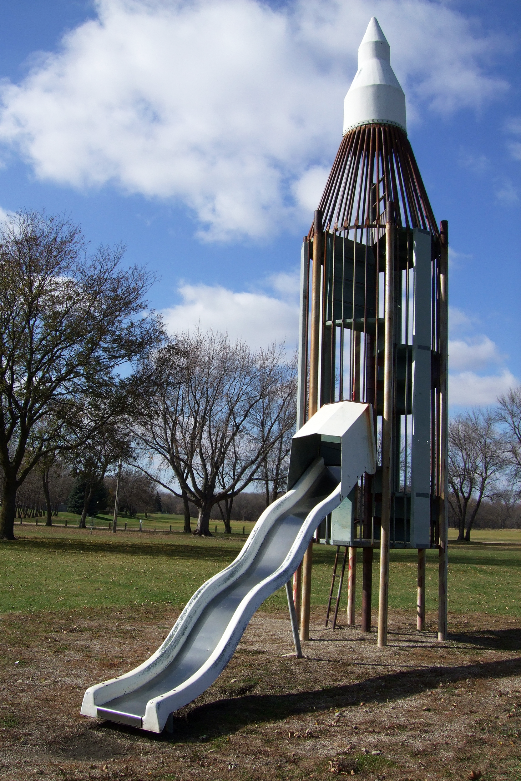 Metal and wooden rocket climber with a metal slide on a playground, large bare tress in the background with a bright blue sky with white clouds