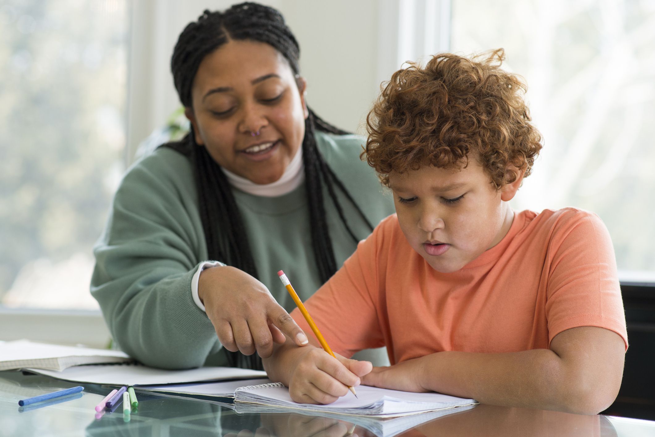Mother smiling gently while helping her young son with homework at home, both are sitting at a glass top table, he is holding a pen on paper, with a blurred background of the wall and two windows showing a sunny day and trees