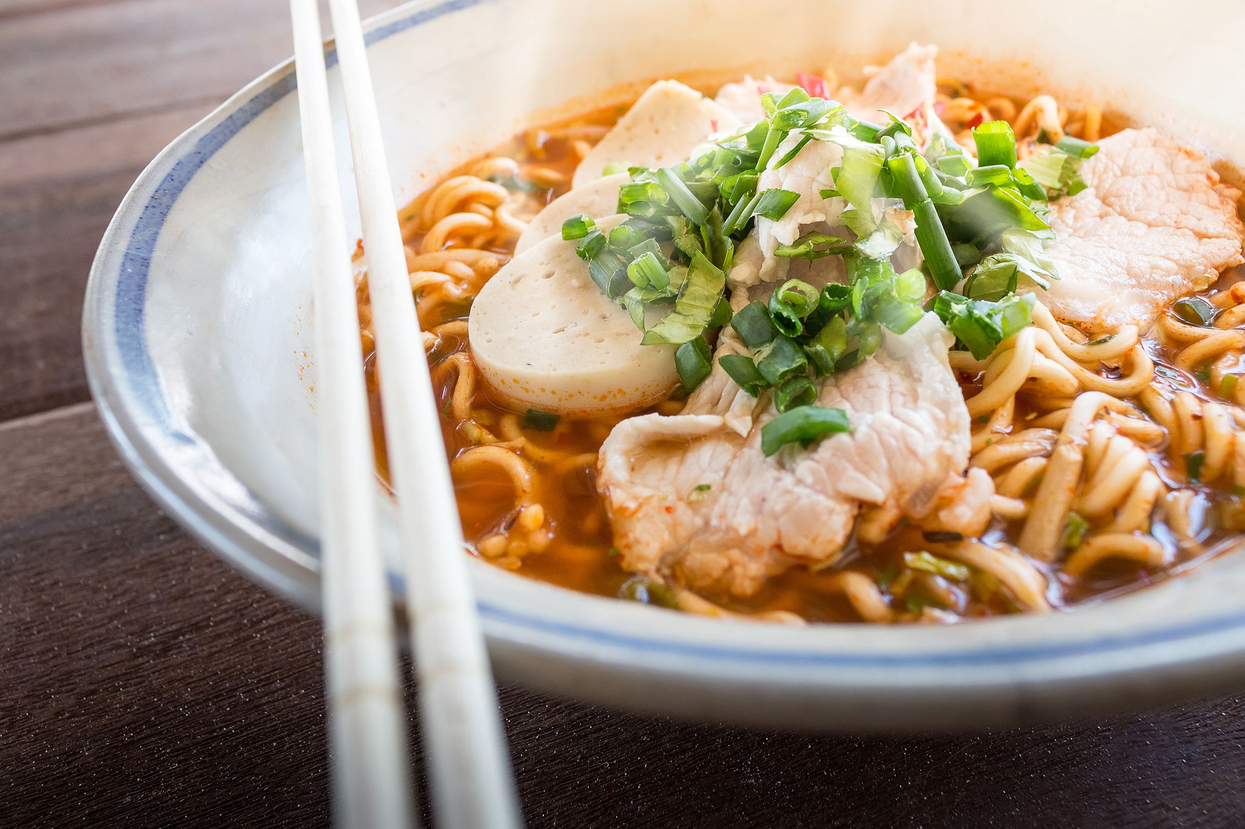 Ramen noodles with chicken and chives in a blue-rimmed ceramic bowl with a pair of chopsticks rested around the edge of the bowl on a dark brown wooden table