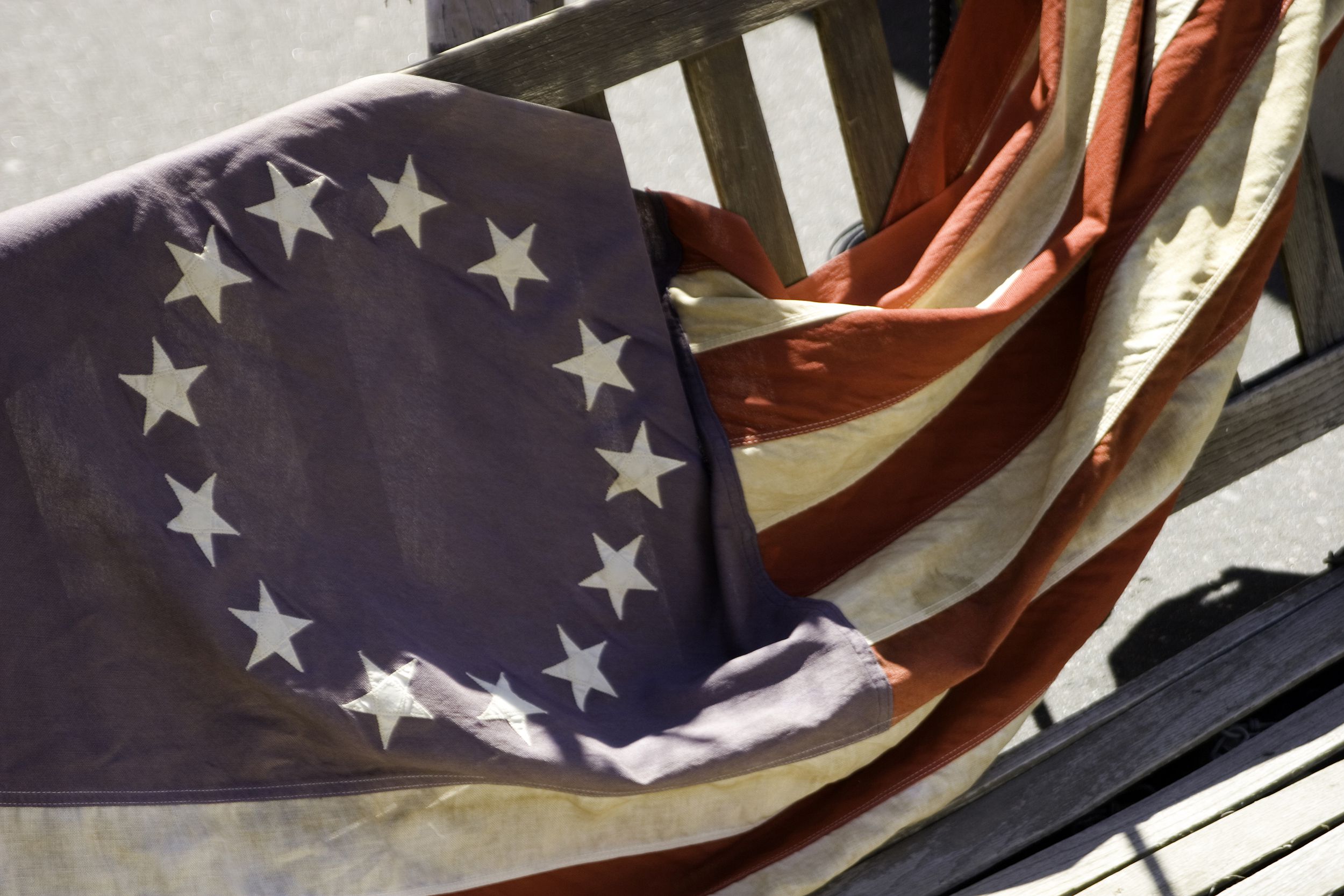 Replica of the original 13 colonies U.S. Flag laying on a wooden bench outside in bright sunlight, with a blurred background of a gravel road