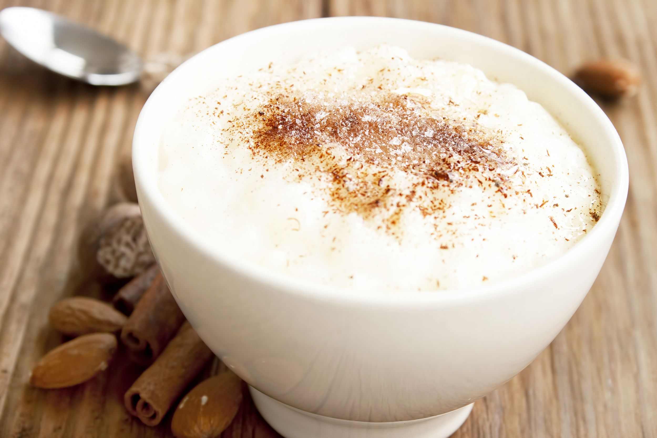 Rice porridge with cinnamon in a white bowl on a wooden table, surrounded by sticks of cinnamon and almonds with a blurred background of a spoon