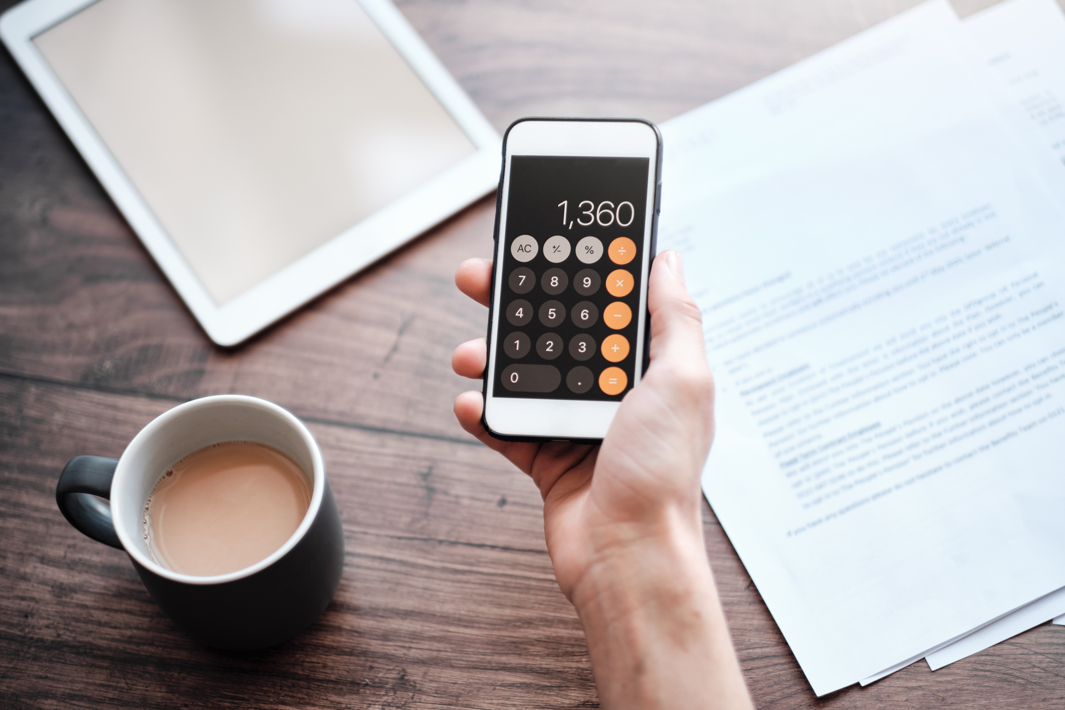Right hand holding a calculator app on a smartphone, middle of the image, against a rustic wooden table with a coffee cup of coffee, a tablet, and documents