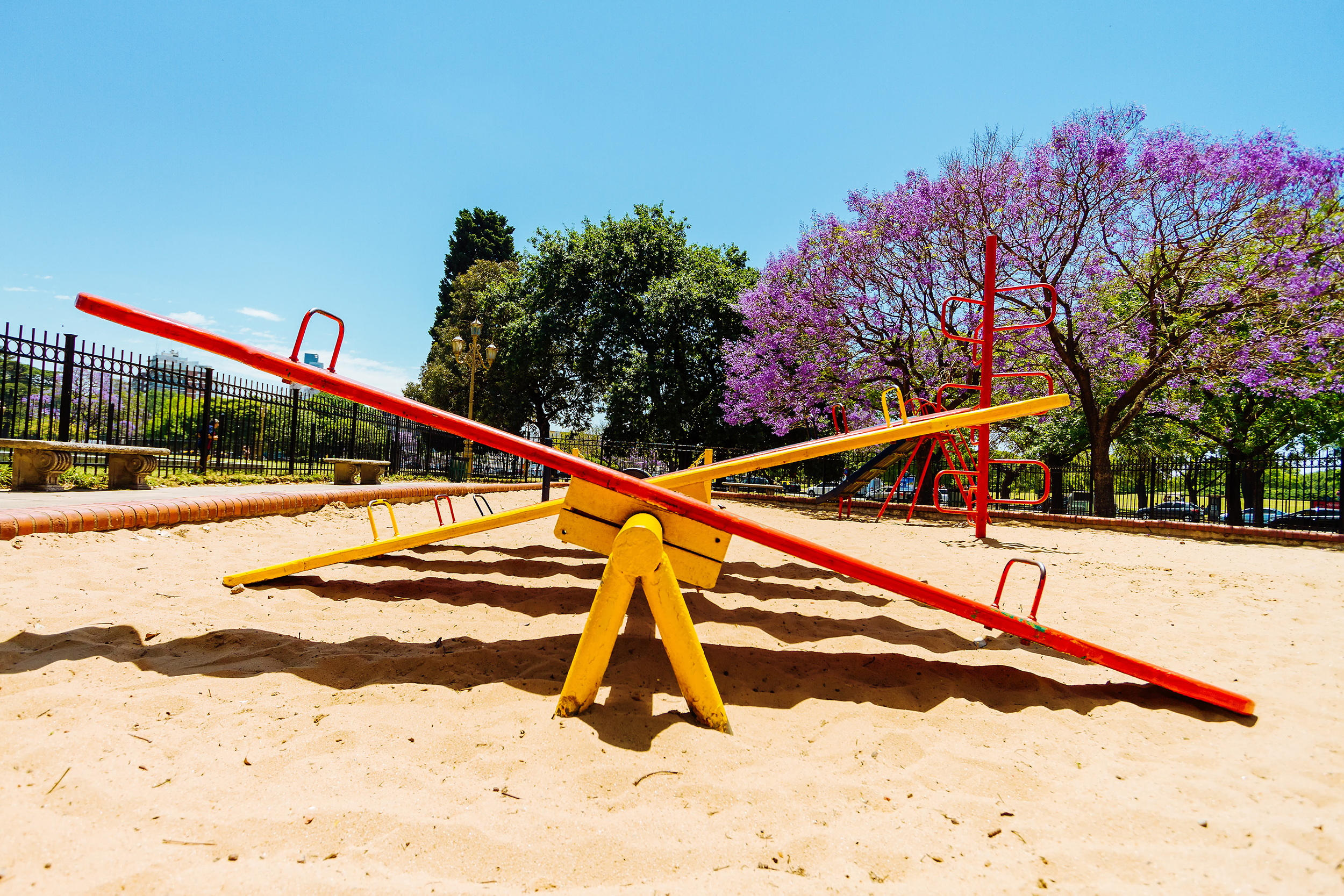 A row of yellow and red seesaws as viewed from the side in the sandbox of a park, a red climbing playground toy on the right in the distance, trees, plants, and a blue sky in the background in summer